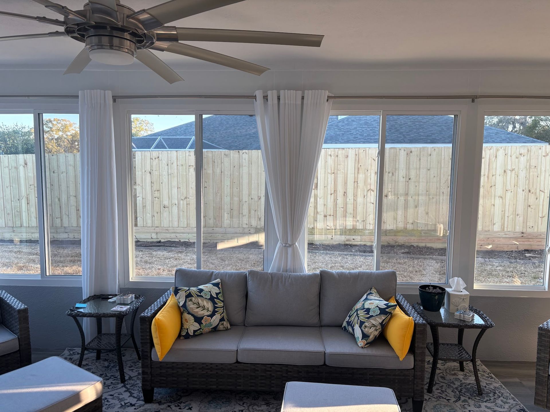 Living room with a gray couch, floor-to-ceiling windows, and a ceiling fan overlooking a fenced yard