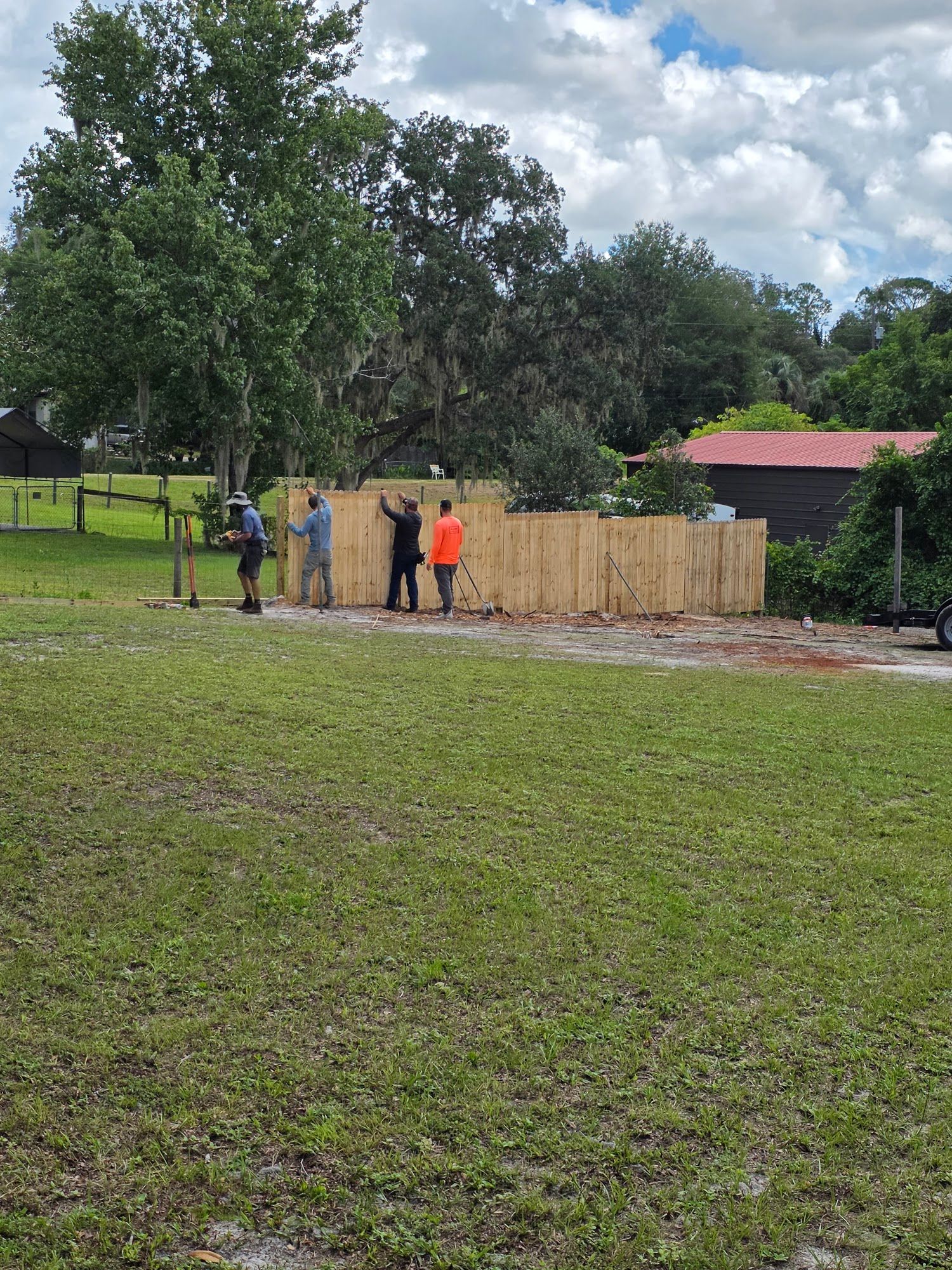 People standing by a wooden fence in a grassy yard under trees, with a red-roofed house in the background