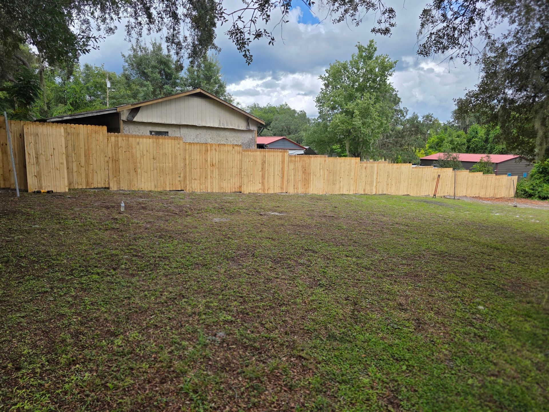 Backyard with a wooden fence, grassy yard, trees, and a small house under a partly cloudy sky
