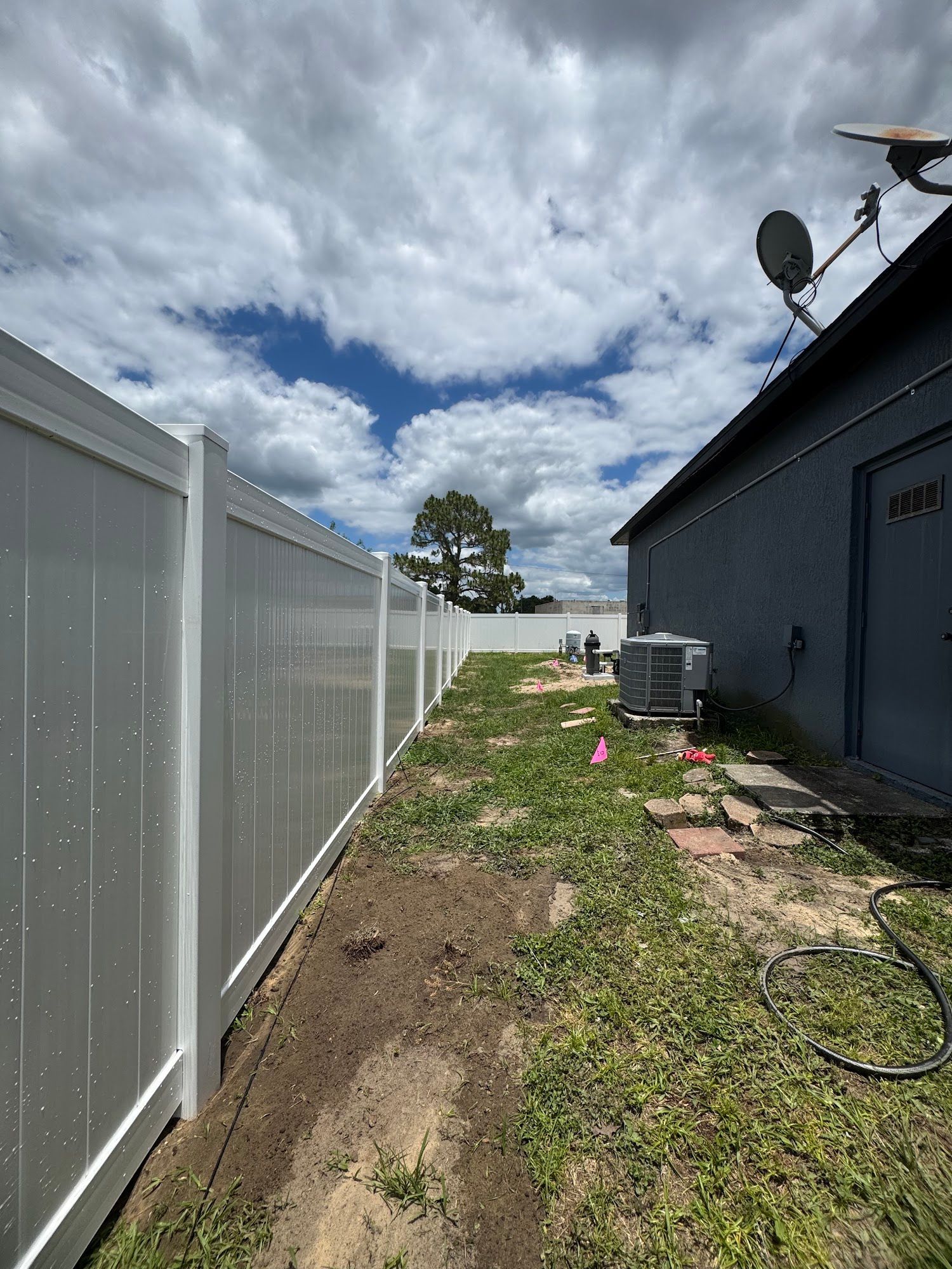 Narrow side yard with white fence, blue house, grass, AC unit, and cloudy sky.