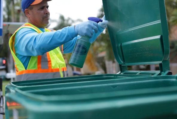 A man is spraying a green trash can with a spray bottle.