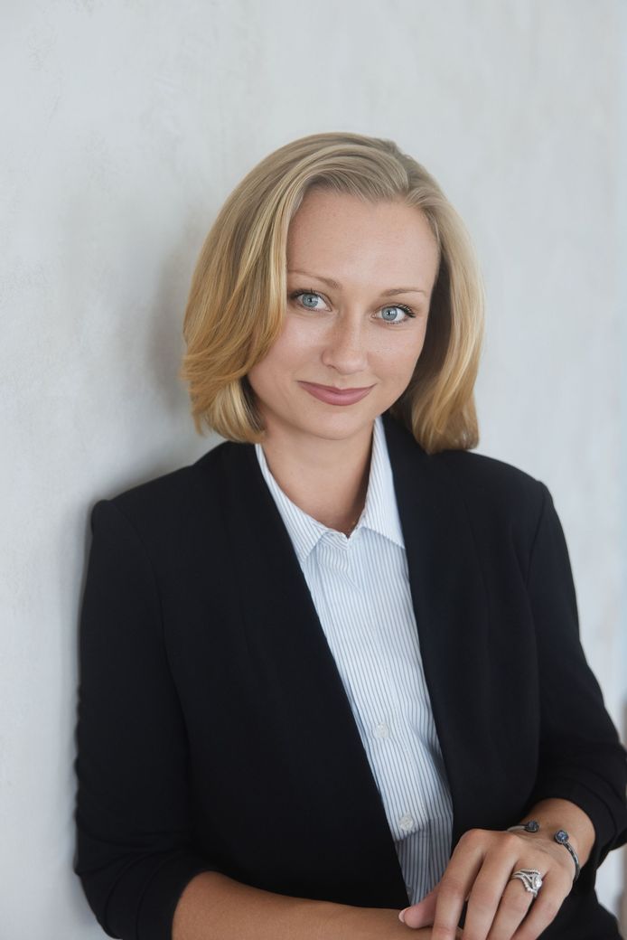 Portrait of Alieta in a black blazer and white shirt, standing against a plain light background.