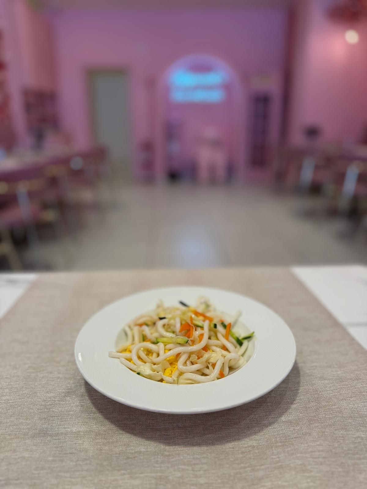 Piatto bianco di noodles e verdure su un tavolo in una sala da pranzo illuminata di rosa