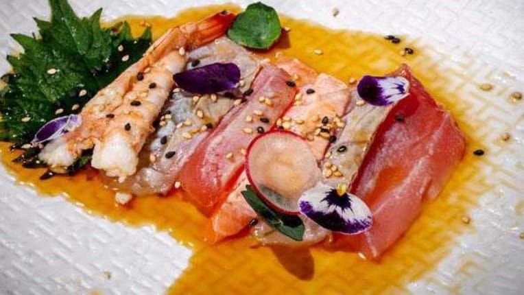 Sashimi di pesce crudo affettato, servito con salsa all’arancia, erbe aromatiche e fiori commestibili su un piatto bianco dalla superficie lavorata
