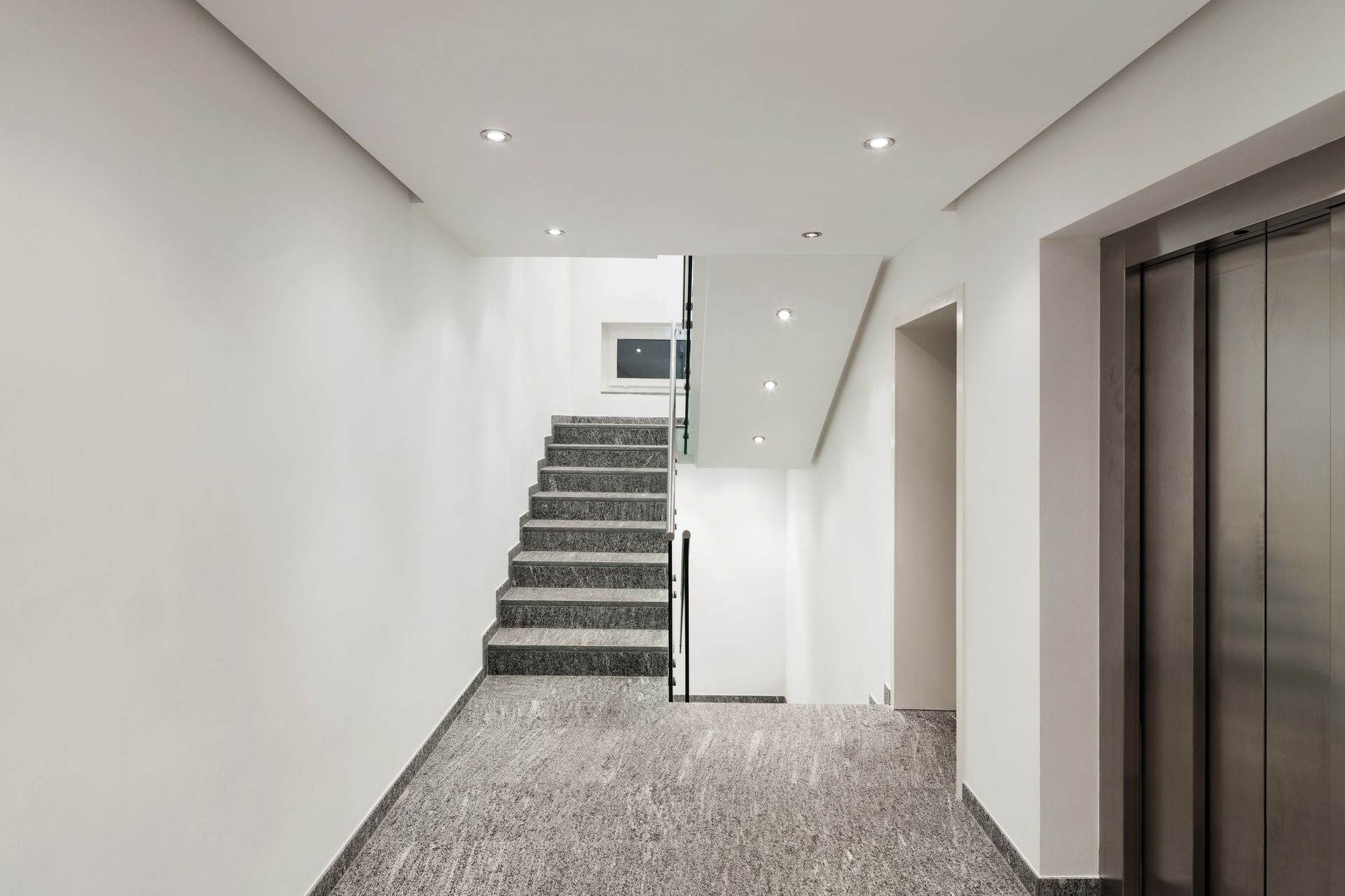 Hallway with granite stairs and elevator on the right. White walls, recessed lighting, speckled flooring. Hallway with granite stairs and elevator on the right. White walls, recessed lighting, speckled flooring.