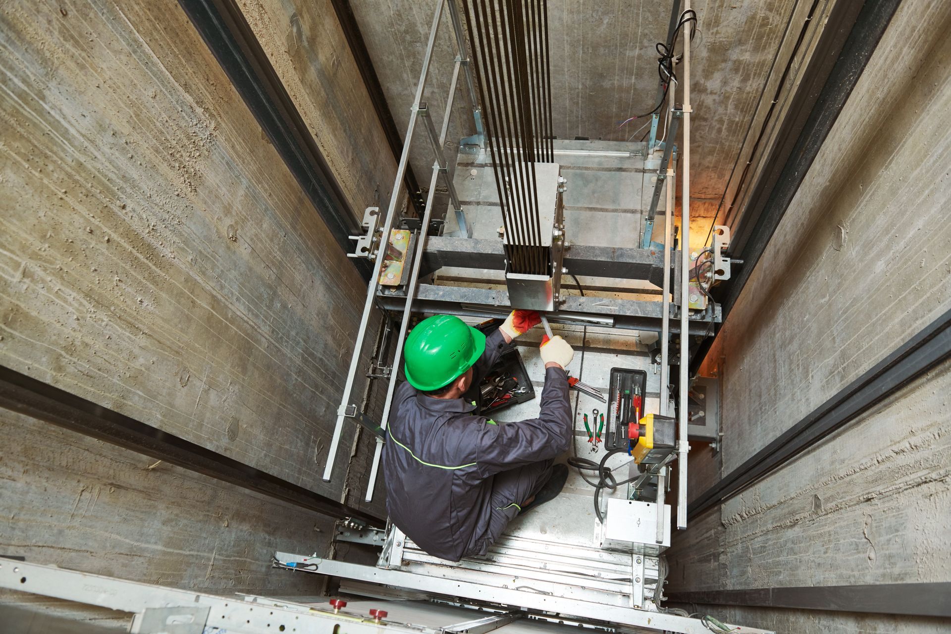 Elevator repair technician in green hard hat, working inside an elevator shaft, using tools.
