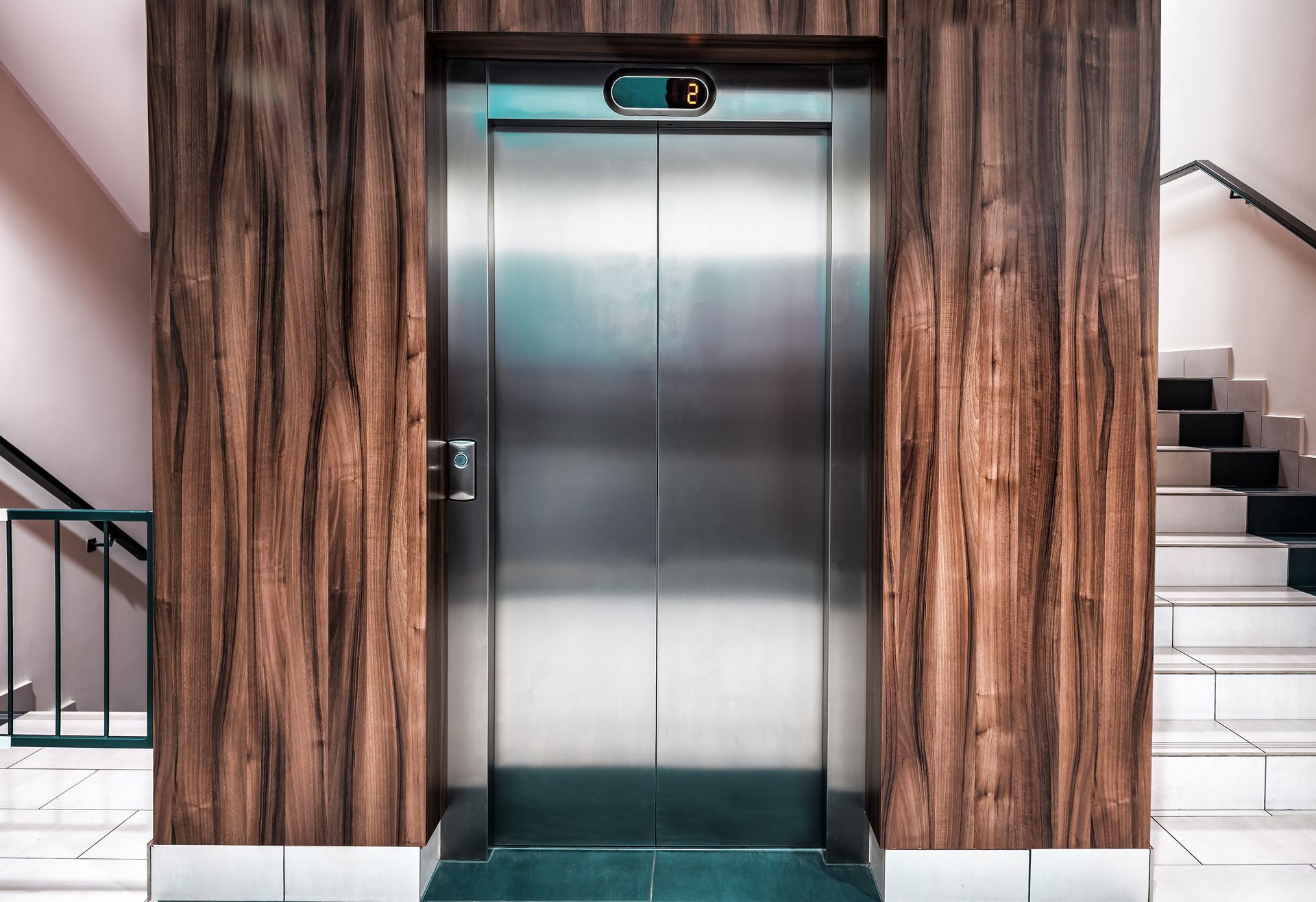 Stainless steel elevator door in a wood-paneled alcove, with stairs and a railing visible. Stainless steel elevator door in a wood-paneled alcove, with stairs and a railing visible.