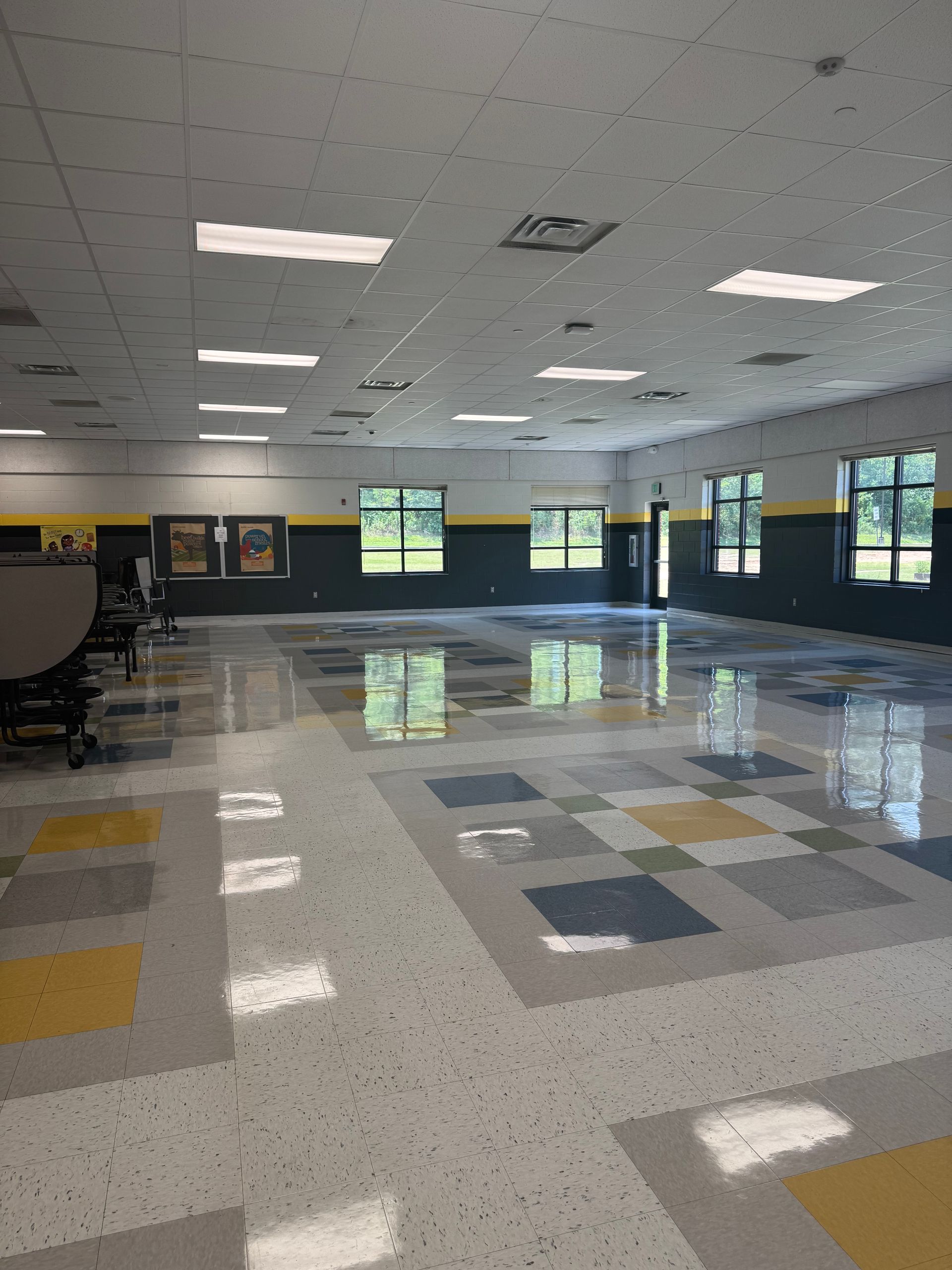 Empty school cafeteria with reflective floor and colorful square tile pattern. Windows, grey walls, and ceiling lights.