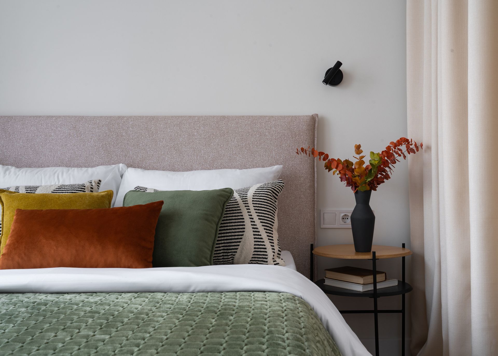 Cozy bedroom corner with a bed, green and rust pillows, bedside table, and vase of orange flowers. Cozy bedroom corner with a bed, green and rust pillows, bedside table, and vase of orange flowers.