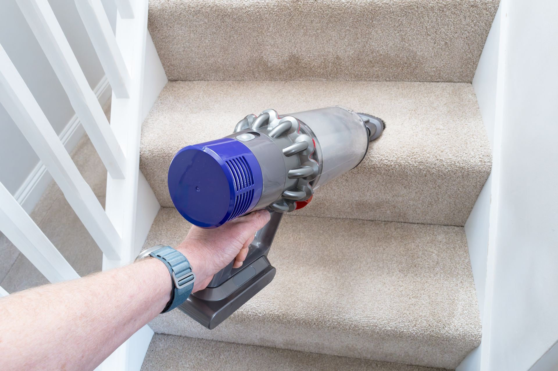 Handheld vacuum cleaner being used on a carpeted staircase near a white railing Handheld vacuum cleaner being used on a carpeted staircase near a white railing