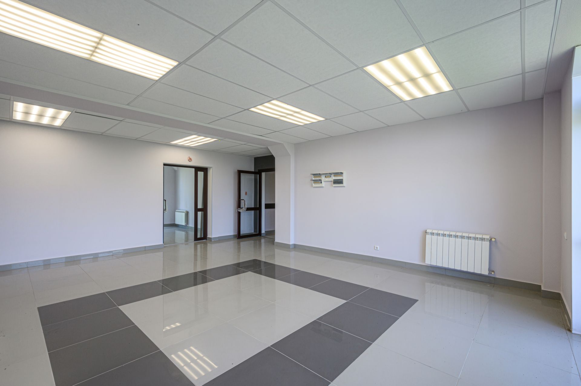 Empty office room with gray tile floor, white walls, ceiling lights, and doorways at the back Empty office room with gray tile floor, white walls, ceiling lights, and doorways at the back