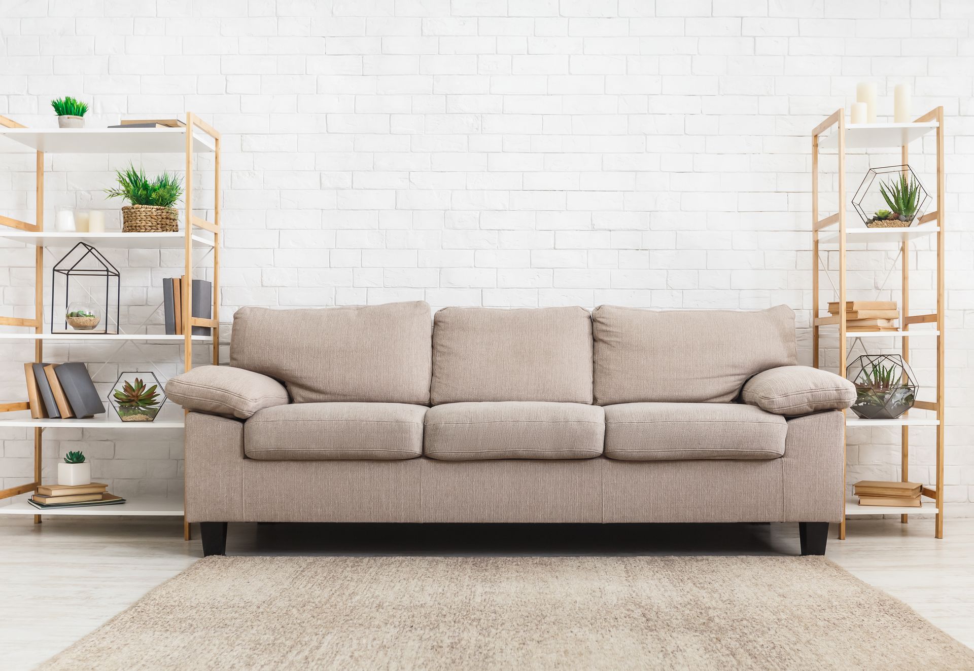 Beige sofa centered in a bright living room with white brick wall and wooden shelves holding plants. Beige sofa centered in a bright living room with white brick wall and wooden shelves holding plants.