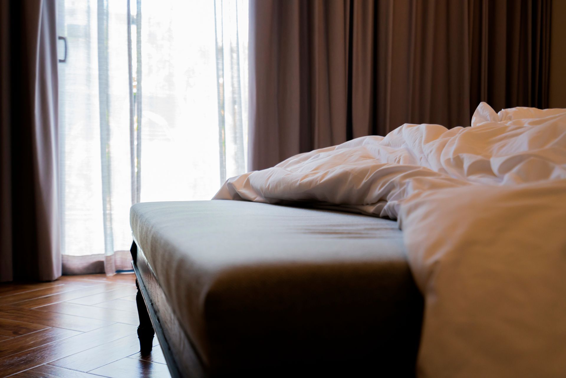 Hotel room bed beside a bright window with curtains and rumpled white bedding Hotel room bed beside a bright window with curtains and rumpled white bedding