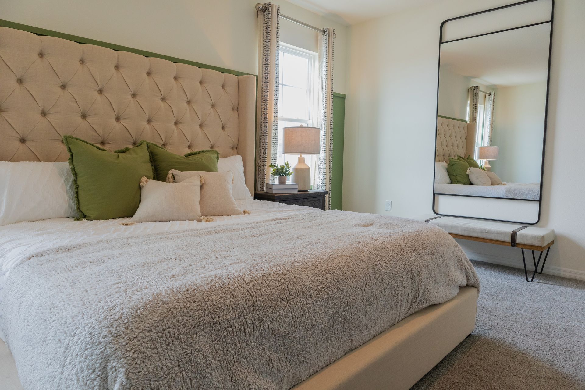 Bright bedroom with tufted beige bed, green accents, full-length mirror, and window light Bright bedroom with tufted beige bed, green accents, full-length mirror, and window light