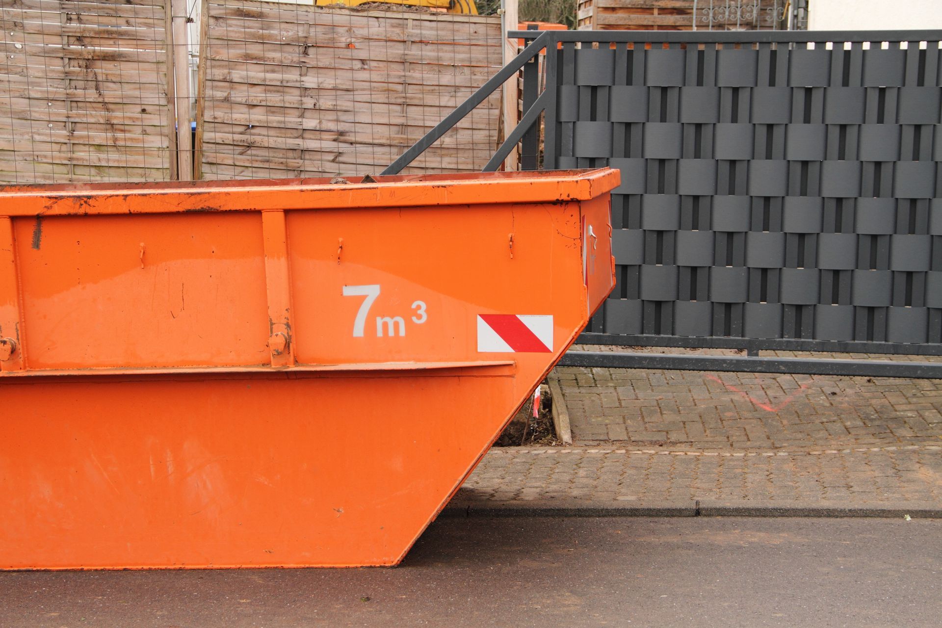 An orange dumpster is parked on the side of the road in front of a fence.