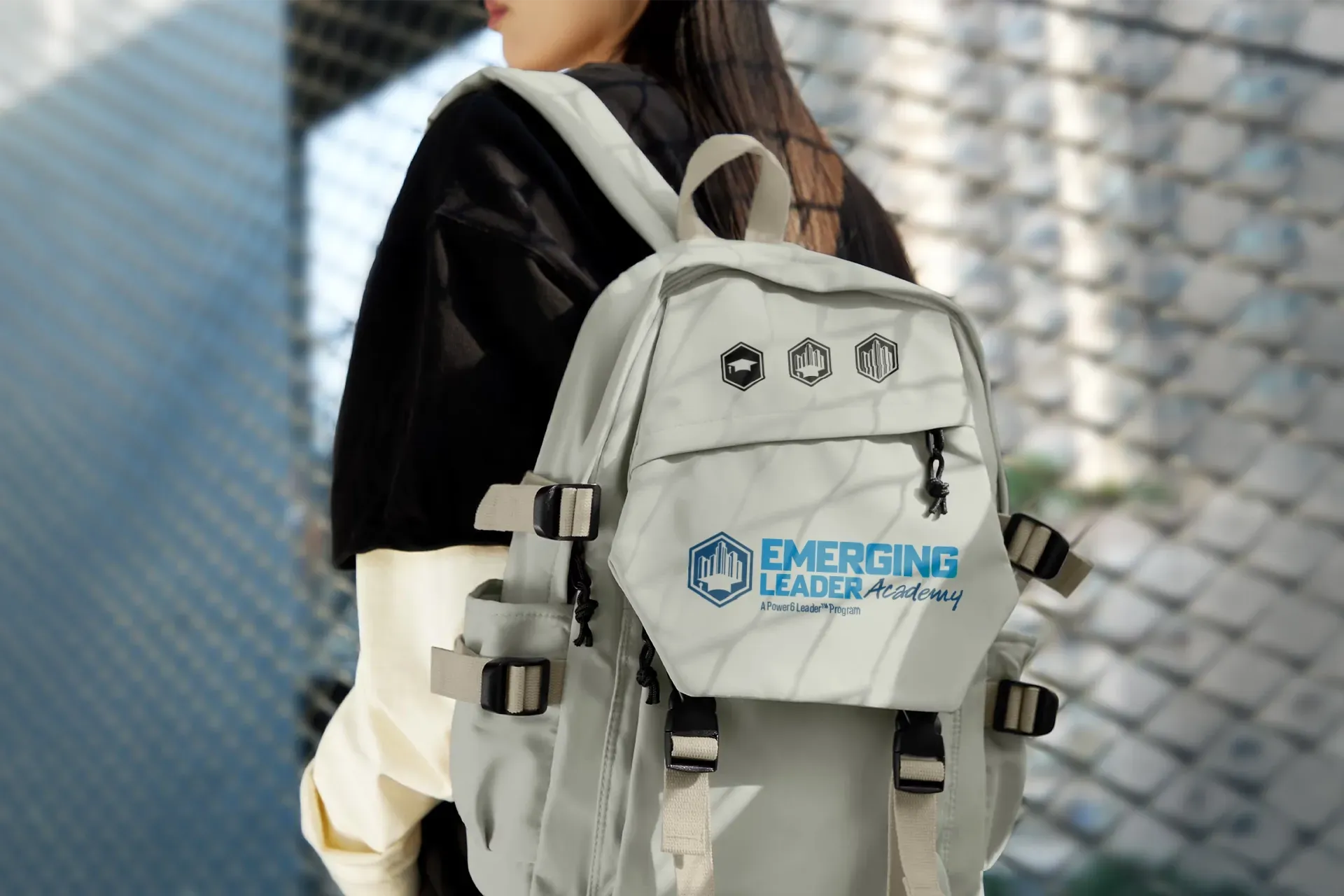 Person wearing a light gray backpack with blue logo, standing outdoors against a blurred fence backdrop