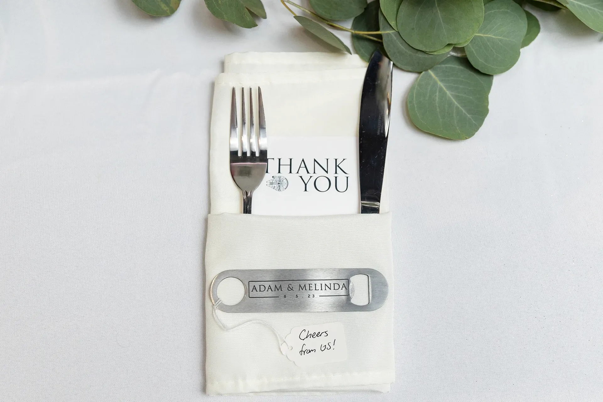 Wedding thank-you favor with fork and knife wrapped in white napkin, topped with greenery