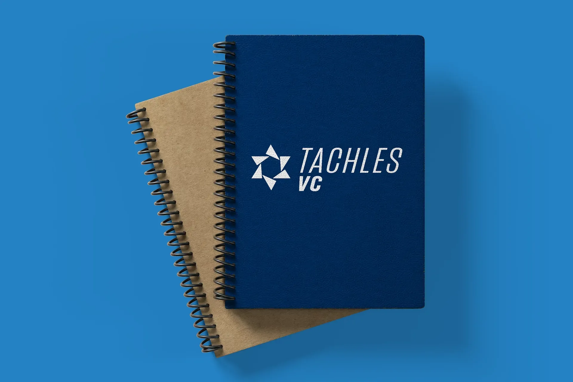 Two spiral notebooks stacked on a blue background, one navy with “TACHLES VC” logo.