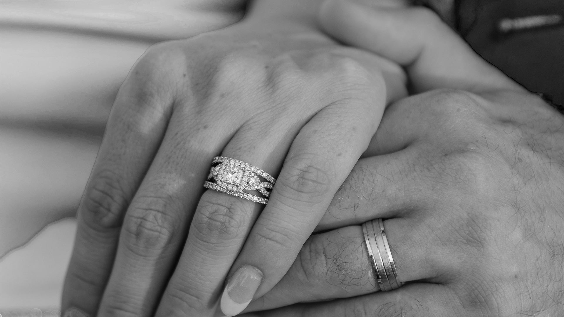 Two hands clasped together, each wearing a ring, in a black-and-white close-up.