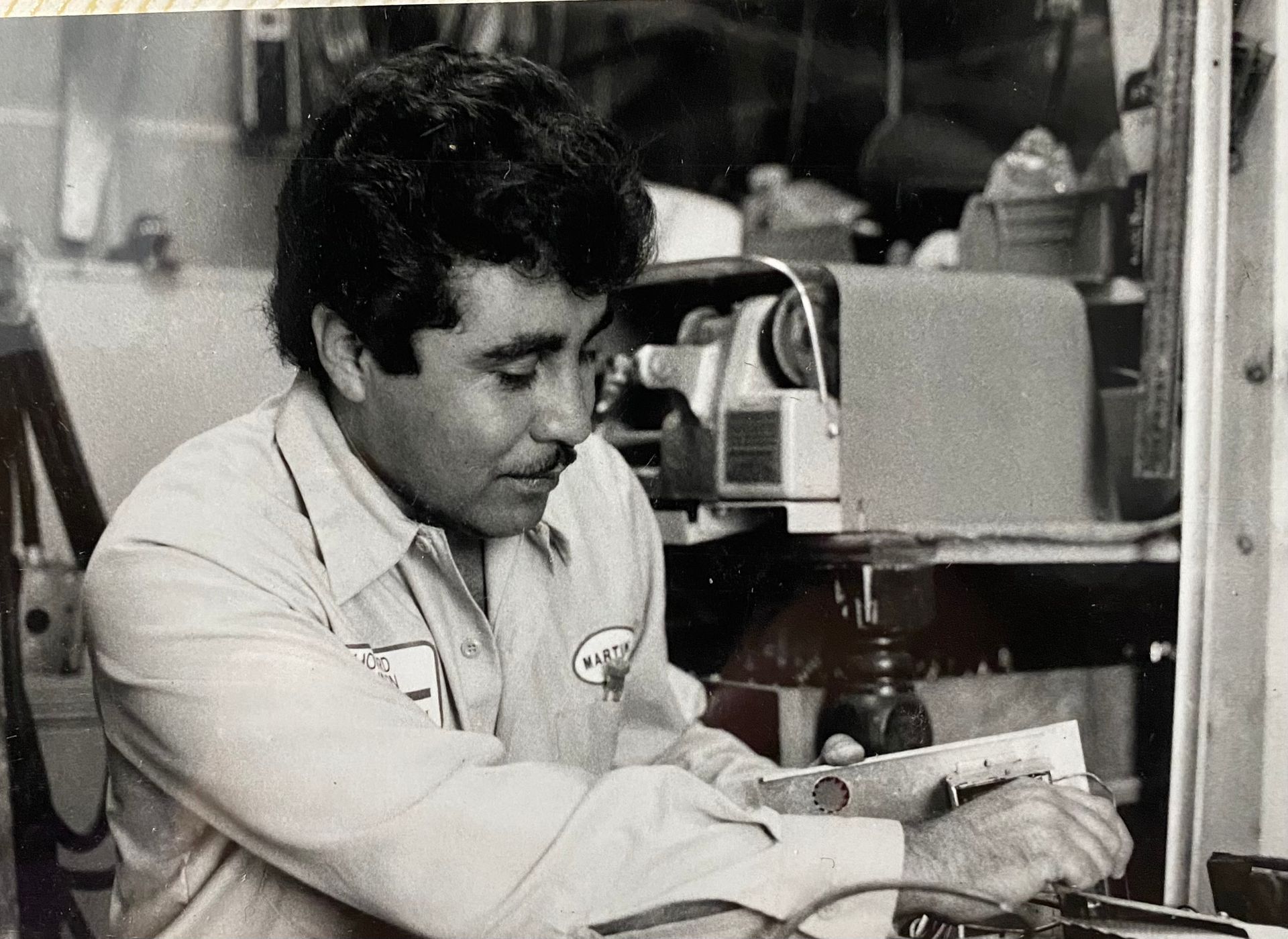 Man working at a desk in a workshop, concentrating on a task beside a machine.