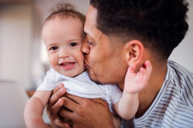 Man kissing smiling baby on the cheek, both in a close-up embrace indoors