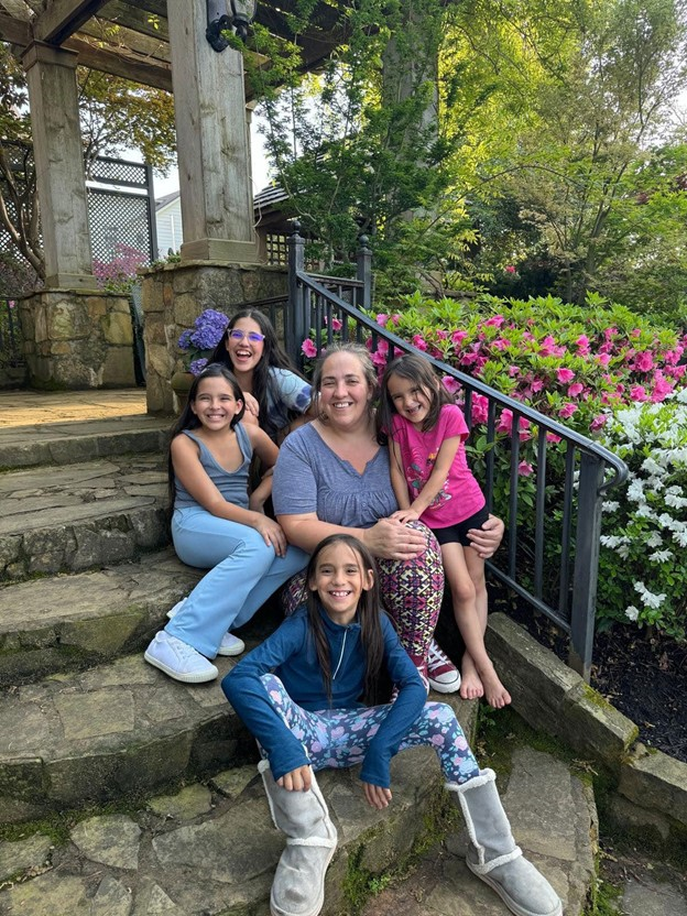Five friends sitting on stone steps outdoors beside a black railing and pink flowers, smiling at the camera.