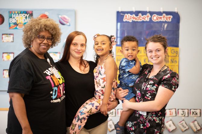 Four people posing in a classroom, with a child being held and a colorful alphabet bulletin board behind them