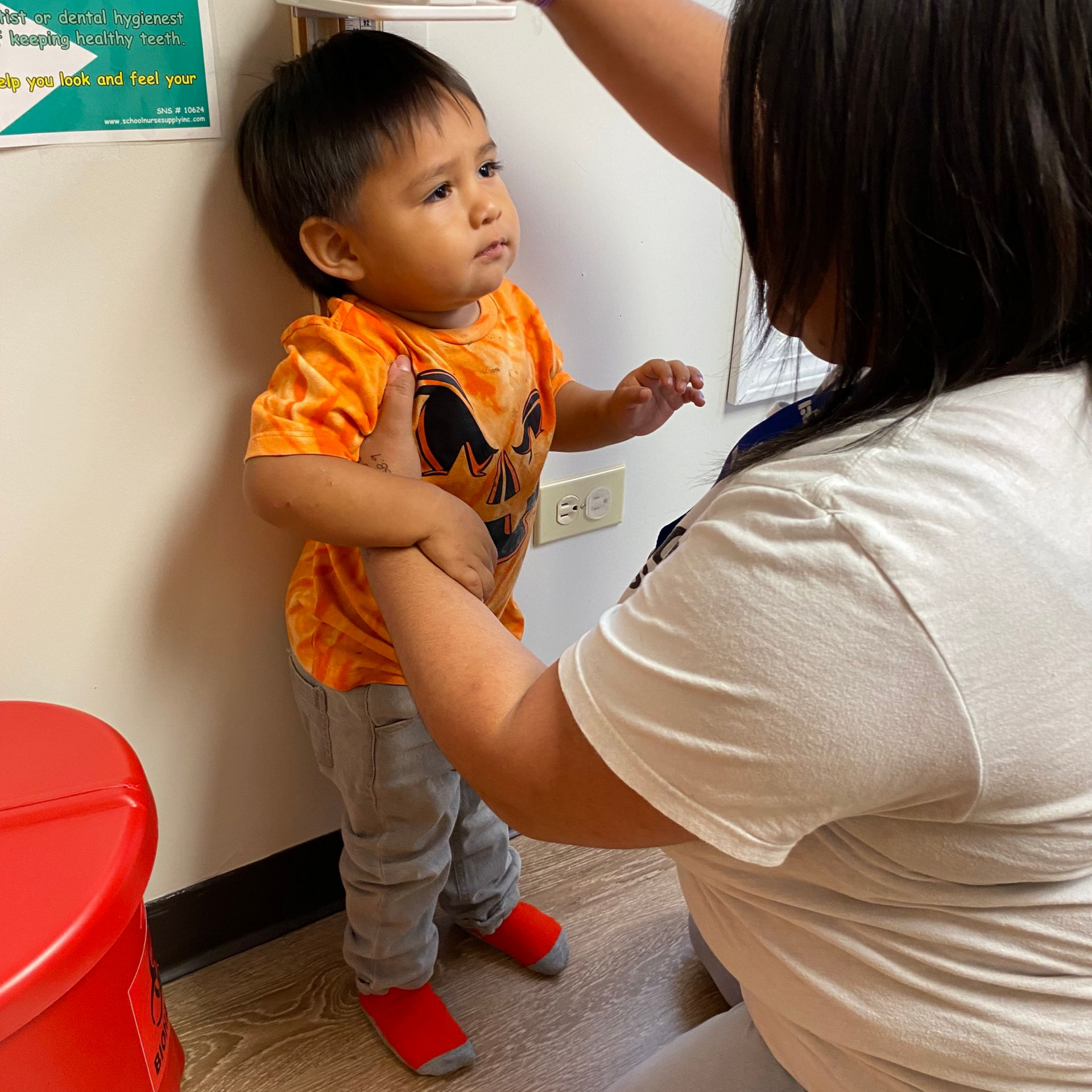 Adult helping a child stand in a clinic room, with a red stool nearby.