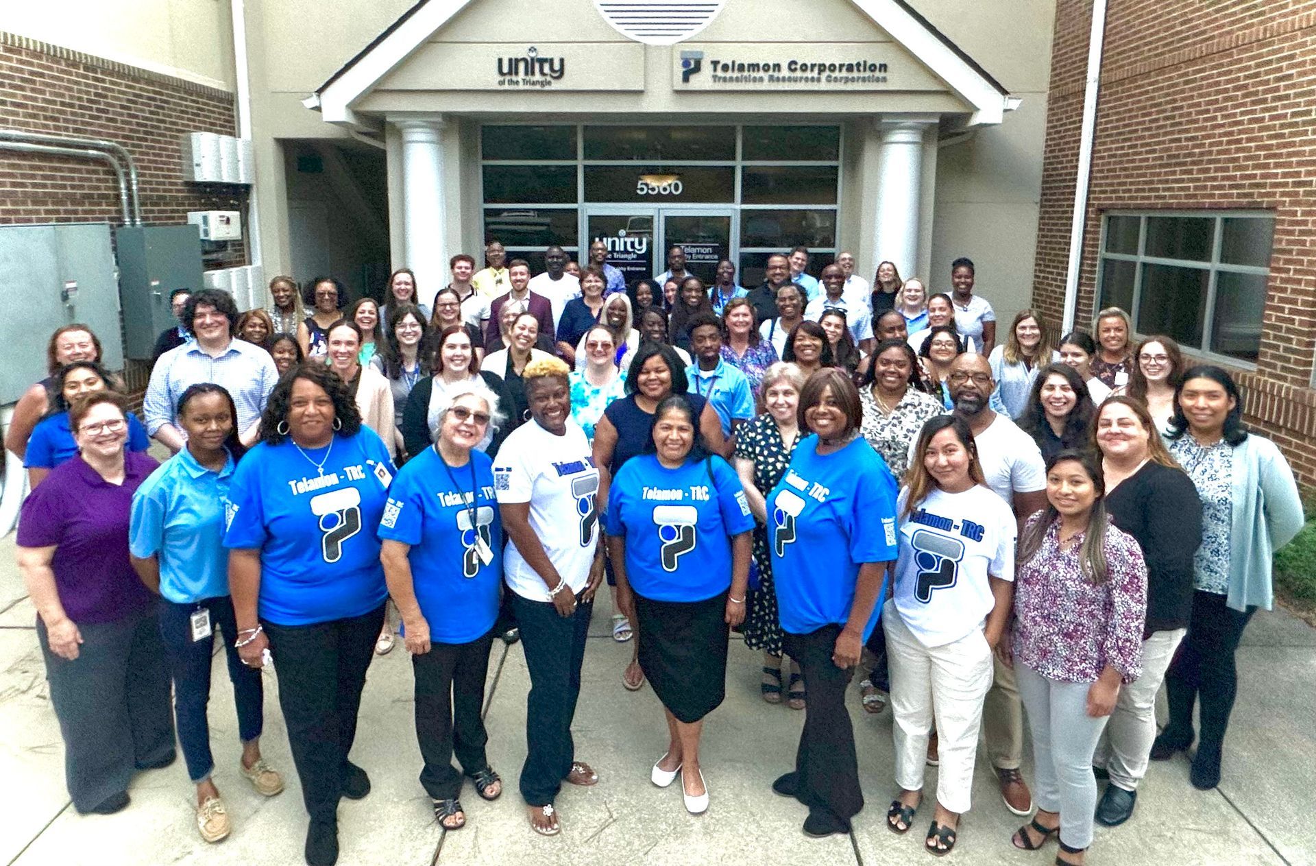 Large group posing outside a building entrance, many in blue T-shirts and smiles.