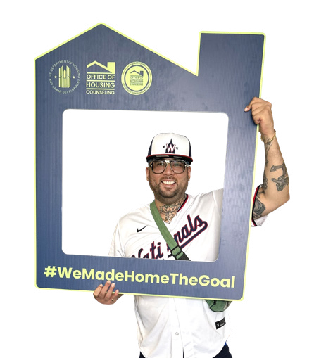 Man holding a blue home-shaped frame with #WeMadeHomeTheGoal, wearing a white baseball uniform and helmet