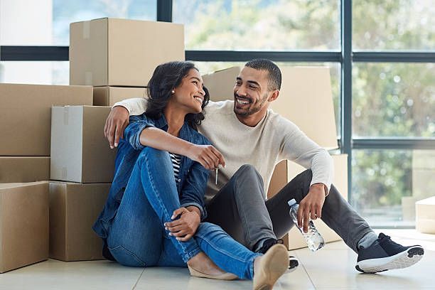 Couple sitting on the floor, smiling and hugging beside stacked moving boxes in a bright room