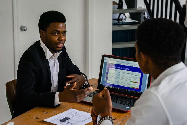Two people in a meeting across a table, with a laptop displaying a spreadsheet and papers spread out.