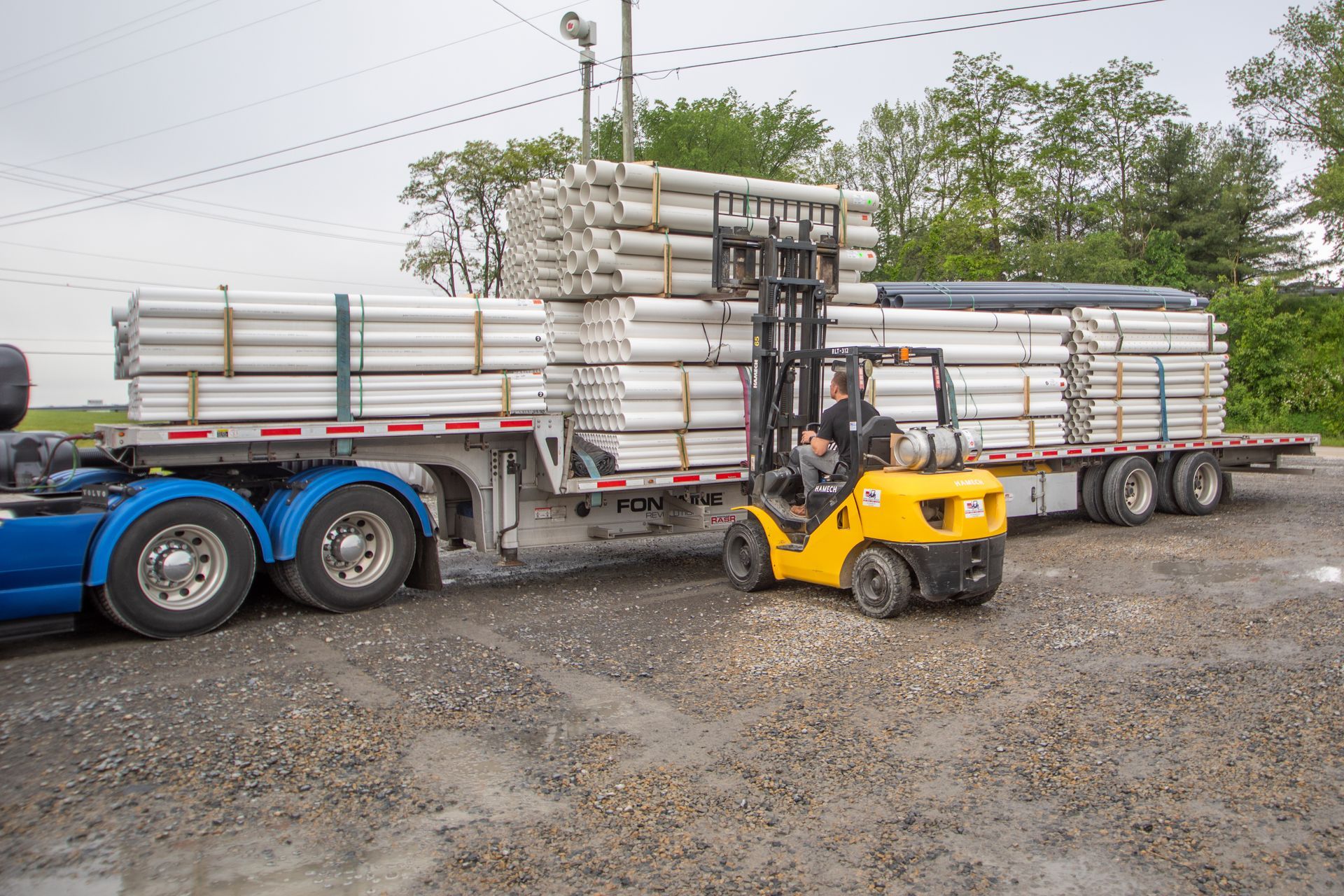 A yellow forklift loads cylindrical pipes onto a blue semi-truck trailer in an outdoor area.
