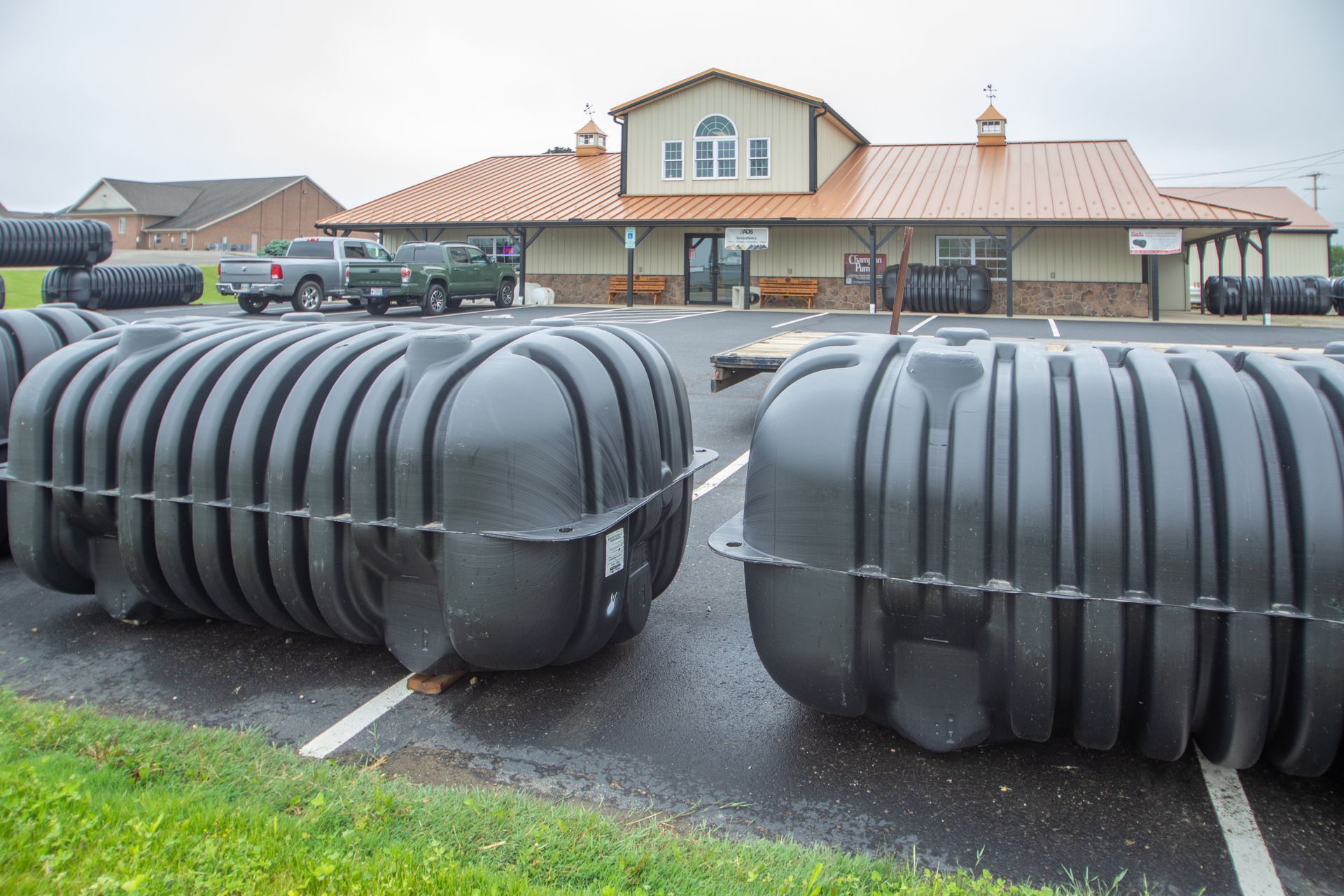 Black plastic septic tanks in a parking lot with a building in the background. Trucks are parked nearby.
