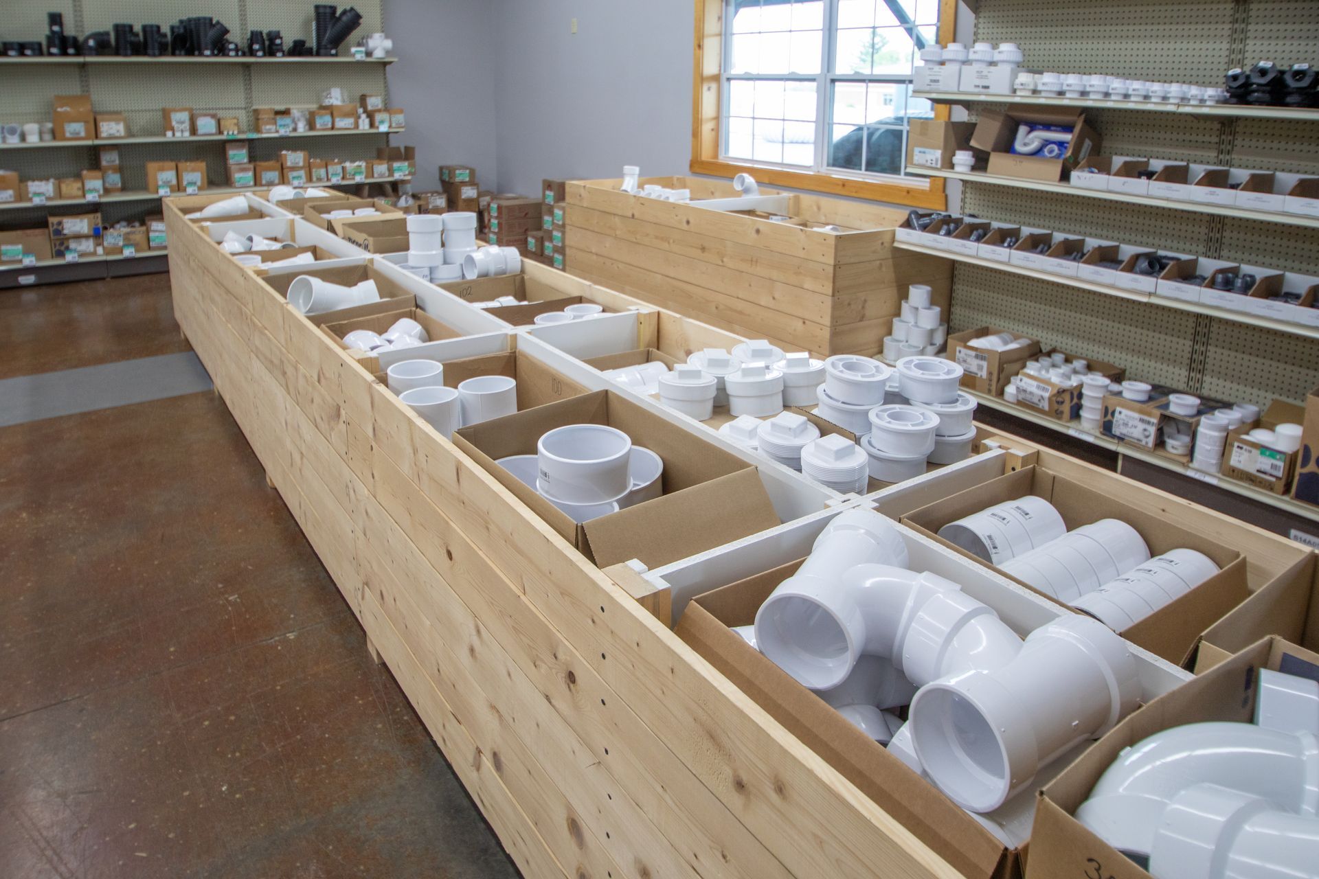 Shelves of white plumbing fittings in a hardware store; wooden bins display various shapes, sizes.
