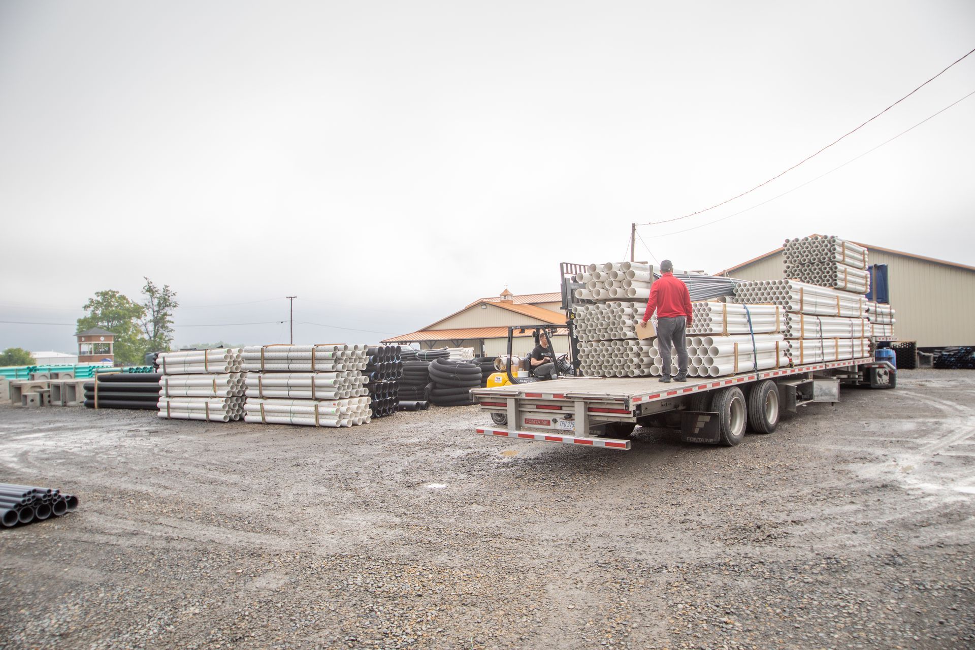 Truck loaded with poles at a supply yard. A person in red stands on the truck bed. Overcast sky.