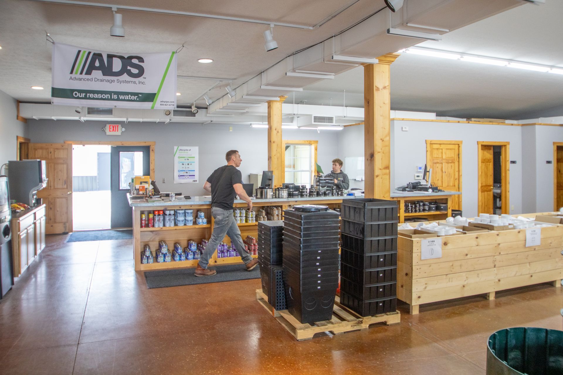 Inside a store: man walks past stacks of black bins. Another person stands at a counter. Display shelves filled.