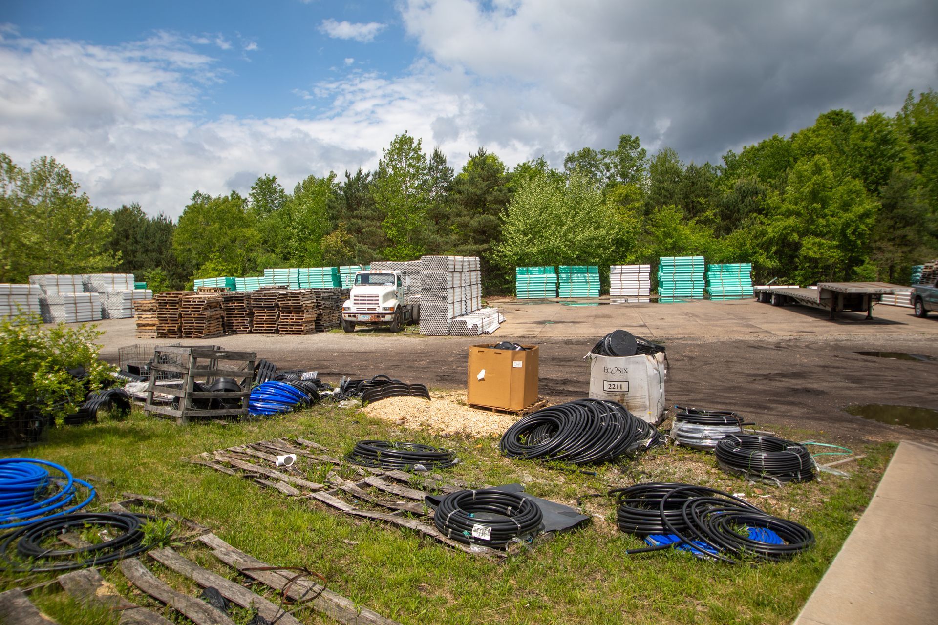 A lumberyard with stacks of building materials, a truck, and scattered equipment, set against a tree line under a cloudy sky.