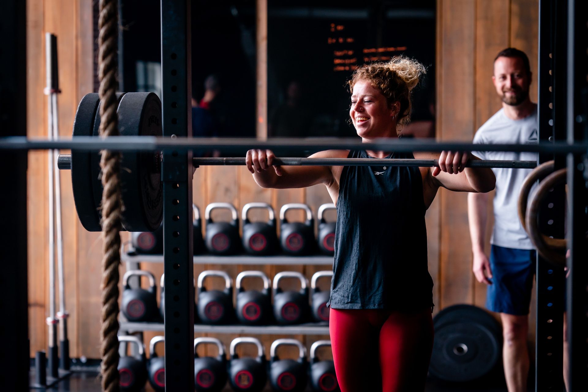 Een vrouw tilt een halter op in een sportschool, geholpen door een man; ze lacht