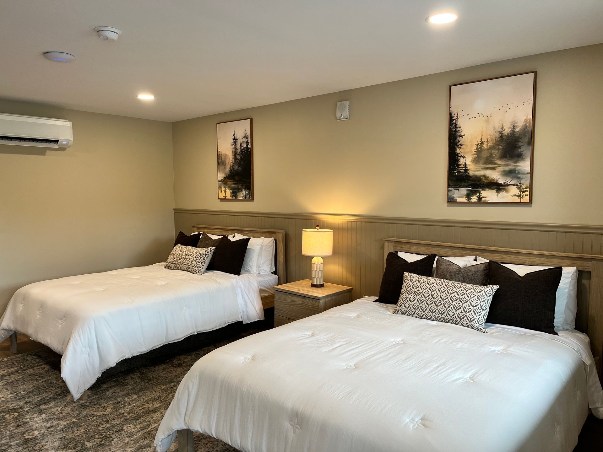 Bright bedroom with a wooden bed, bedside lamps, woven chair, rug, and framed landscape art