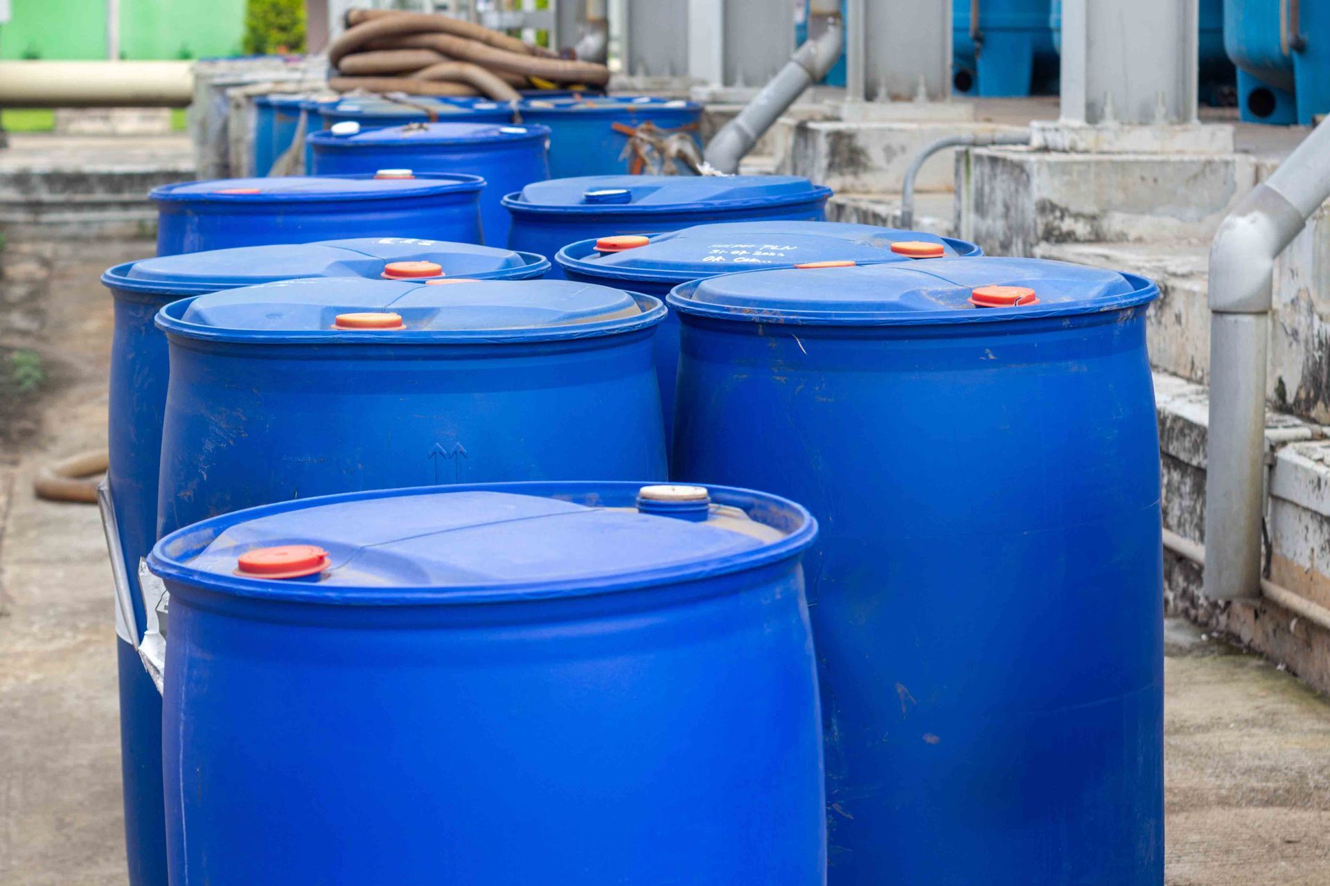 Blue industrial barrels lined up in a factory yard, with pipes and equipment in the background