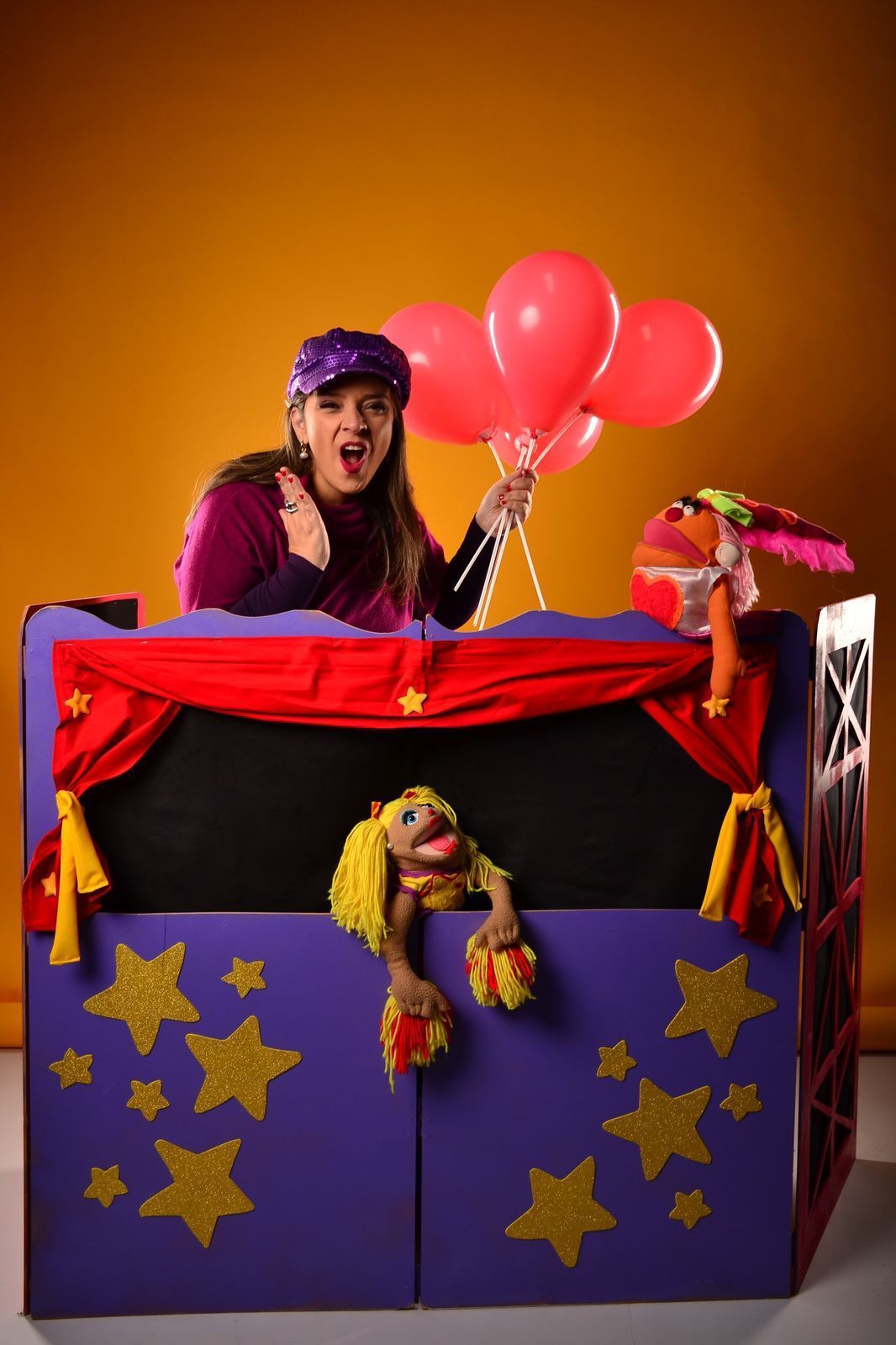 Persona que sostiene globos rosas, realizando un espectáculo de marionetas desde un escenario morado con estrellas amarillas.