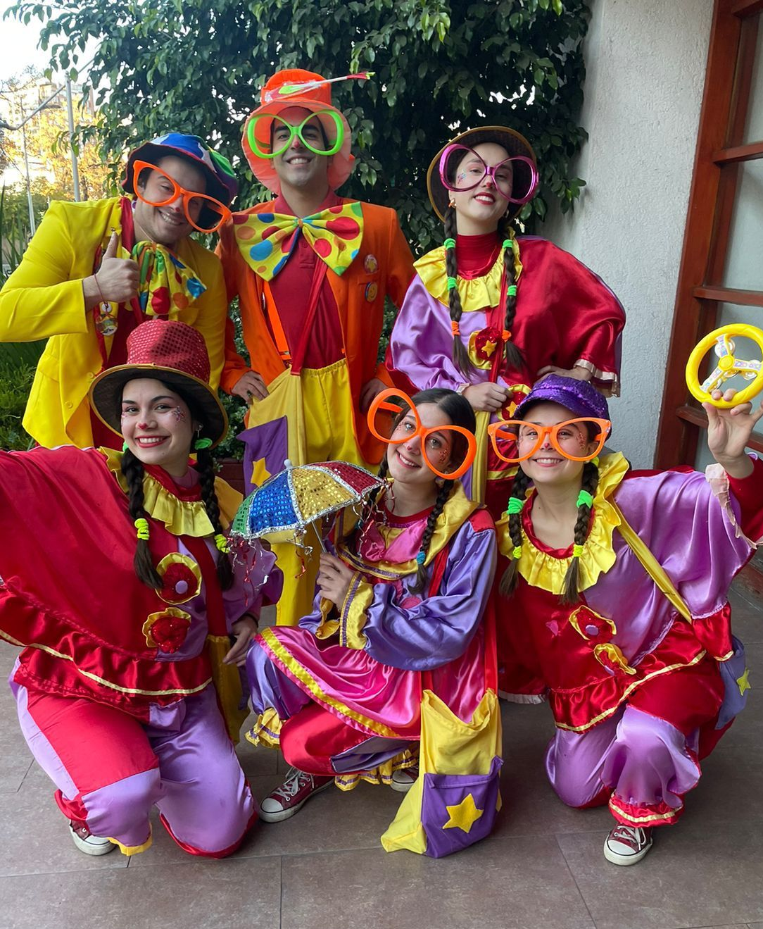 Grupo de siete payasos con trajes coloridos y gafas naranjas, posando al aire libre.