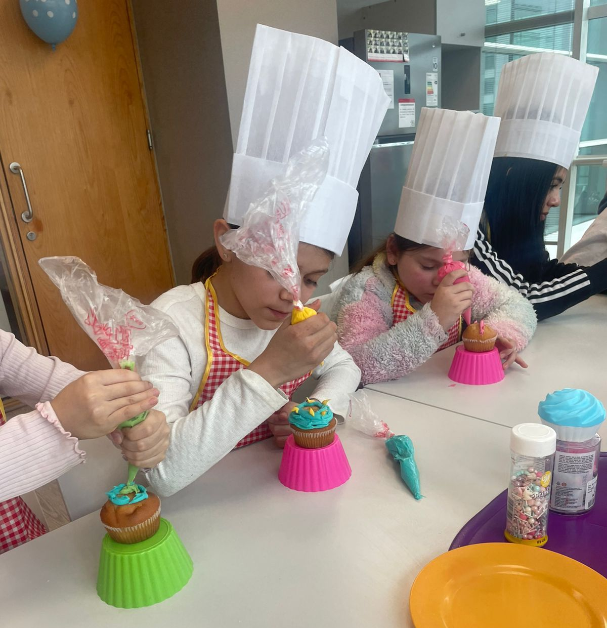 Niños con gorros de chef decorando cupcakes en un mini taller de chef. Borde de arcoíris colorido.