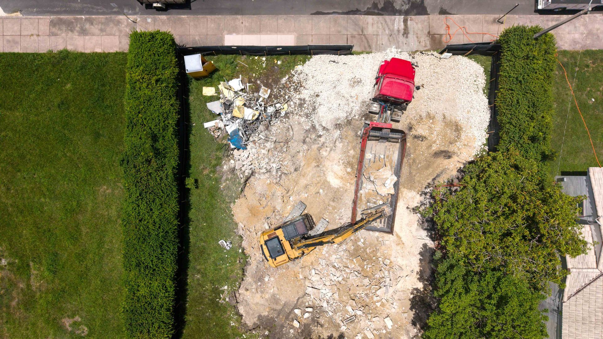 Aerial view of a small excavator digging in a yard beside trimmed hedges and a sidewalk.
