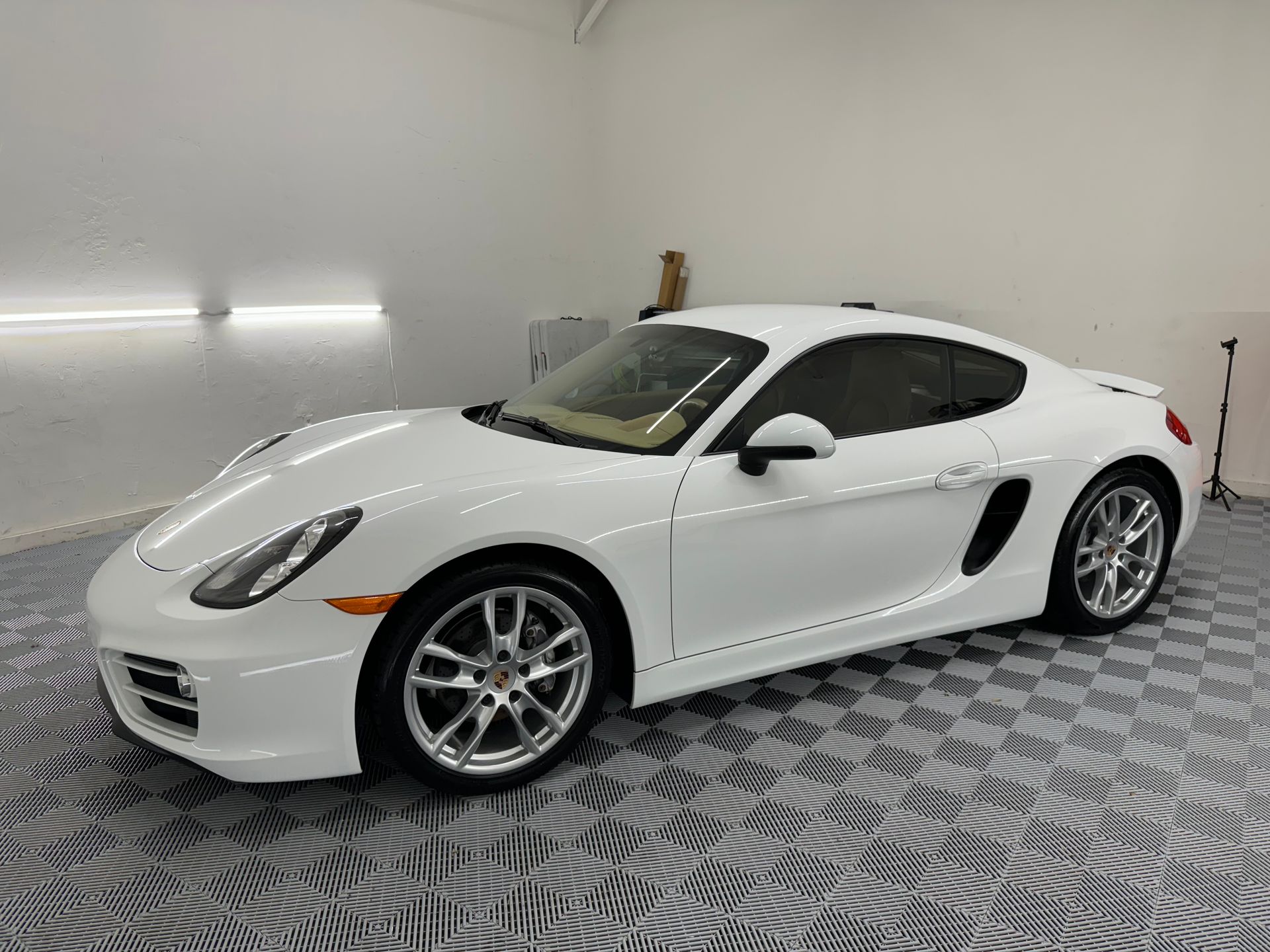 White Porsche Cayman coupe, parked indoors on a checkered floor.