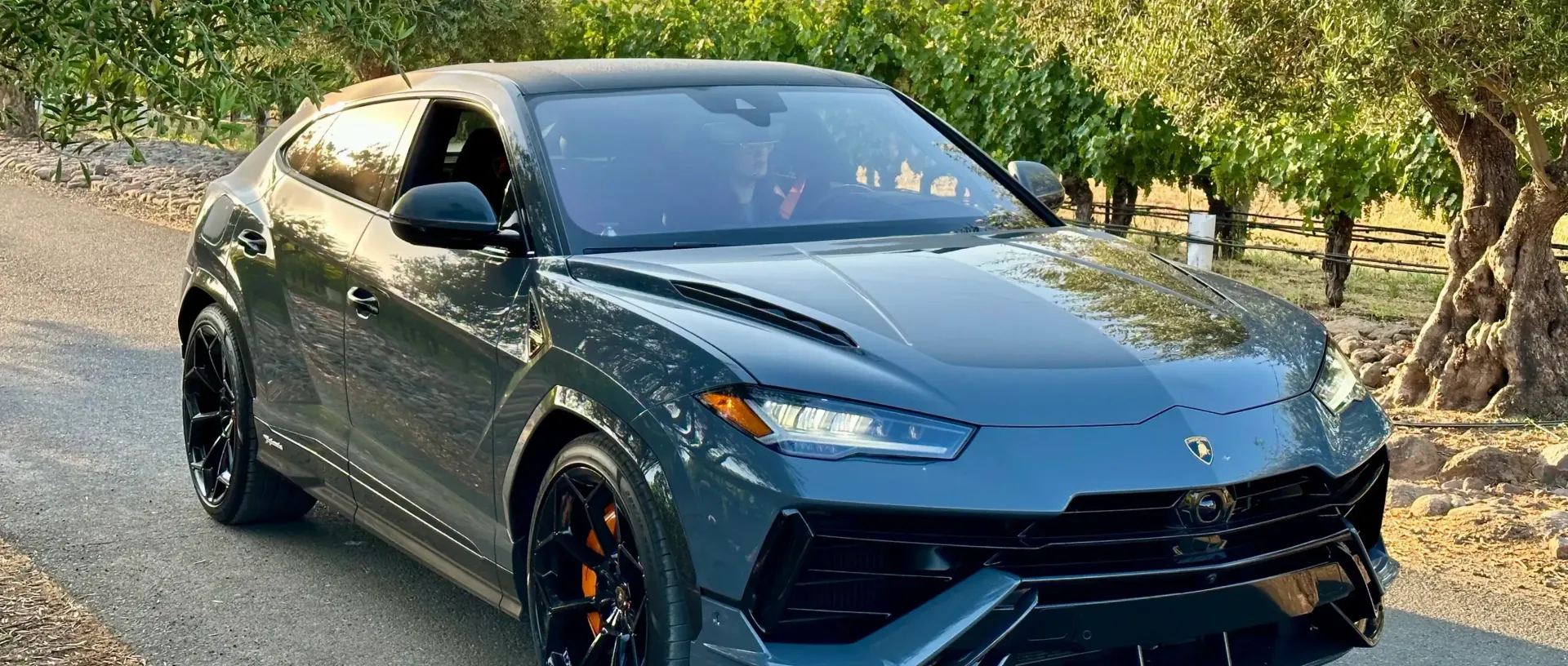 A gray Lamborghini Urus SUV on a road with trees in the background.