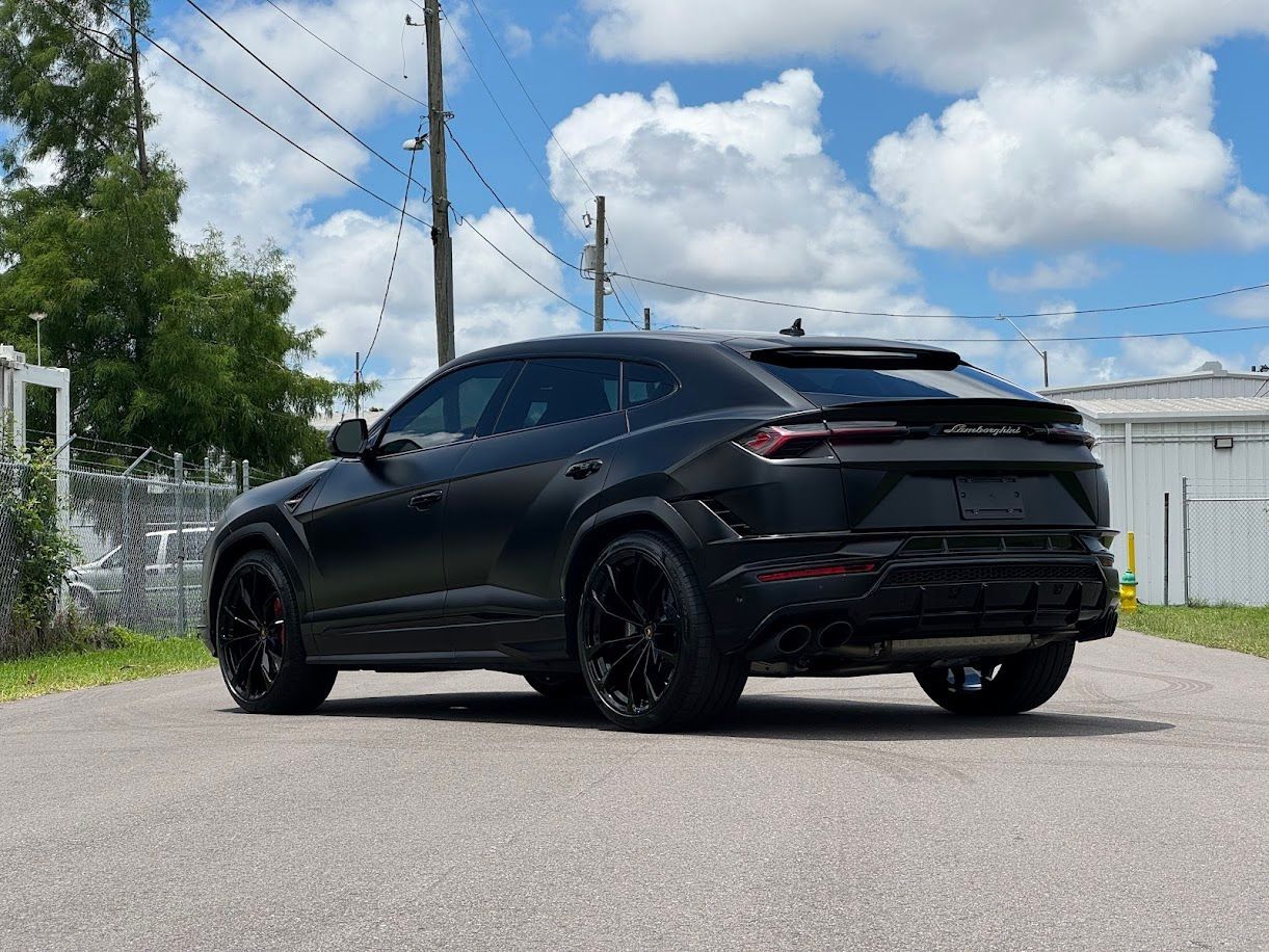 Black Lamborghini Urus SUV on a road, against a cloudy sky.