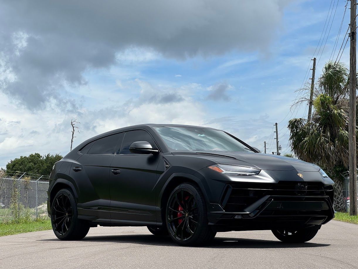 Black Lamborghini Urus SUV parked on a paved road, under a cloudy sky.