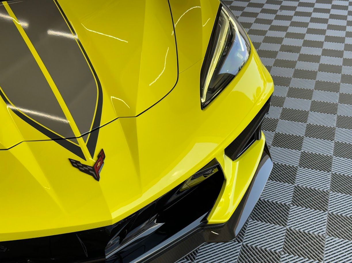 Bright yellow Chevrolet Corvette with black racing stripes on the hood, parked indoors.